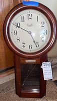 Front view of vintage Versailles wall clock showing wood frame, clock face with Arabic numerals, and lower glass door.