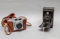 Both cameras side by side, showing Kodak Hawkeye folding camera on right and Balda camera with brown leather case on left.