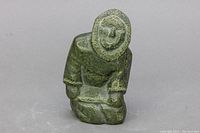 Front view of Inuit soapstone carving showing kneeling figure with detailed facial features and hooded traditional clothing.