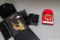 Three jewelry items shown: two gold-tone Joan Rivers necklaces displayed in a black case and a black multi-beaded necklace in a black box, with a red box holding a KJL hinged bracelet next to them.