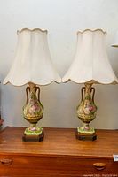 Pair of vintage floral hand-painted porcelain table lamps with scalloped fabric shades on a wooden surface against a white wall.