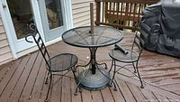 Full view of iron mesh bistro table with two chairs on wooden deck; shows umbrella pole and cover folded on chair