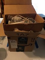 Two stacked cardboard boxes containing various clock parts including gears, rods, and clock faces, packed loose and in bags.