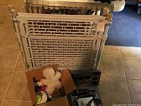 White plastic dog gate with metal connectors, metal pet pen enclosure behind it, box with assorted dog toys including a white plush toy, and black Andis dog hair clippers case with accessories.