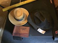 Photo of black Dynatec Texas hat, straw hat with blue and brown band, and leather pilot log cover placed on a black surface.
