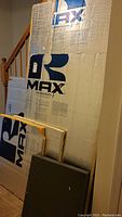 Four handmade insulation panels, two large R-MAX branded foam insulation panels with yellow foam edges and foil facing, leaning against a wall with a staircase in background.