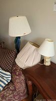 Photo showing floor lamp with colorful twisted glass base and pleated cream lampshade, retro table lamp with hexagonal floral base and white lampshade, and two separate cream lampshades on a couch.