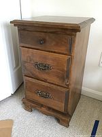 Front and side angle of wood nightstand showing three drawers with one knob and two metal handles, wood grain, and carved feet.
