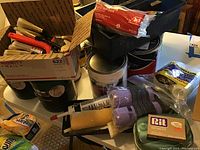 Photo showing a box of assorted paint brushes, paint rollers, a roll of plastic drip cloth, Rit brand caulk, and other painting supplies on table.