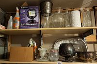 Photo showing shelf with multiple glass vases, spray bottles, a box with lock set, ceramic vase, silverplate bowls and aluminum roasting pan below.