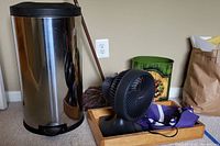 Wide view showing Simplehuman trash can, Holmes Blizzard fan, mop handle, green decorative waste bin, wooden box with fabrics, and folded flag.