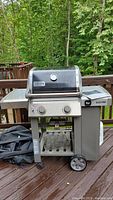 Front view of Weber Genesis II gas grill on wooden deck with manual and grill cover nearby. Grill has two control knobs and a lid thermometer. Grill is wet from rain.