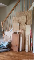 Pile of assorted wood planks and plywood sheets stacked upright by staircase, showing size and variety.