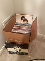 Cardboard box filled with vinyl record sleeves; visible top cover shows male artist portrait over female.