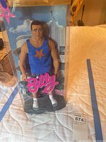 Front view of the Billy doll in its original clear plastic box with visible blue tank top and shorts and pink 'Billy' logo on packaging.