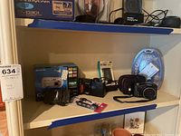 Various electronics including boxed Sony Cyber-shot DSC-P100 camera, Nikon Coolpix 950, Canon PowerShot SX150 cameras, USB hubs, and K40 car radio on shelf.