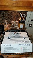 Kitchen counter showing boxed All-Clad 10" petite braiser, wooden knife block set, wooden cutter blocks, and pepper shaker.