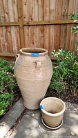 Two large clay pots standing outdoors near a wooden fence. One large pot is upright with visible handles and textured horizontal lines, smaller pot placed in front of it.