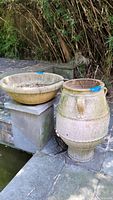 Photo showing two pots: a large round shallow bowl and a tall decorative clay urn with patina and dirt from outdoor conditions.