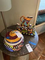 Photo showing the colorful ceramic server with lid and the four-piece tea set including pitcher, sugar bowl, creamer, and tray on top of a round table.