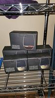 Five black boxes of Riedel wine tumblers arranged on metal shelving, three are for red wine and two for white wine, two tumblers per box.