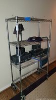 Front view of the chrome wire rack with adjustable shelves, caster wheels, some boxes and a lamp placed on shelves for scale.