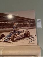Mounted color photo of a racecar number 12 with driver standing on it at Indianapolis Motor Speedway, showing the signatures and inscription.