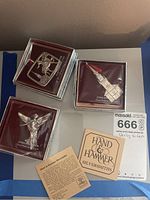 Three sterling silver ornaments shown in clear display boxes with a deep maroon background, including the Public Garden Angel, church steeple, and deer ornaments, along with Hand & Hammer Silversmiths branding cards.