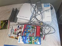 Wii console and accessories laid out on a mattress including console, sensor bar, power adapter, AV cables, stand, remotes, and game cases