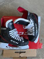 Pair of black CCM hockey skates on top of a red box, white laces tied, viewed from side angle showing overall condition and branding.