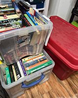 Overview of two transparent plastic containers filled with various books and one large red plastic container closed beside them on wooden floor.