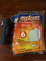 Black Eddie Bauer flashlight lying on wooden surface next to three packages of Hot Shots Toe Warmers still sealed in packaging.