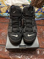 Front view of black Nike Supreme Uptempo sneakers on black shoebox. Showing white Nike swoosh on toe area and strap closure on top of laces.