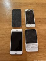 Lot showing four phones: three iPhones including one severely cracked, and one white BlackBerry QWERTY keyboard phone, all on wooden surface.