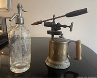 Antique glass seltzer bottle next to vintage metal blowtorch on table.