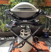 Weber Q grill mounted on folding portable cart with propane tank attached, all-weather wheels visible