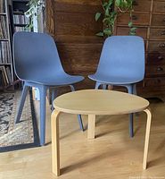 Two blue plastic IKEA outdoor chairs and one small round wooden side table on wood flooring near wood paneled wall.