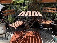Front view of foldable patio table with two foldable wooden slat and metal frame chairs, positioned on balcony with plants in background.