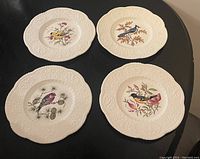Four ceramic plates arranged on black surface showing colorful bird designs and embossed patterns on white backgrounds.
