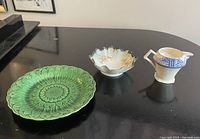 Three china pieces on black surface: green decorative plate, small scalloped bowl with gold trim, and blue & white jug.