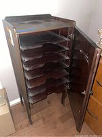 Front view of wooden file cabinet with door open showing seven wave-cut pull-out trays inside.