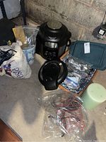 Black Ninja cooking pot with its lid, a basket with assorted silverware, miscellaneous cables and cords, a ceramic utensil holder with additional items, and a large green pillar candle. Cooling racks also visible under plastic wrapping.