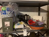 Shelf with various kitchenware and disposable papergoods including plates, napkins, plastic utensils, cups, crock pot, glass container, and serving trays.
