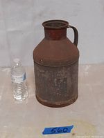 Full view of the antique steel milk jug with a plastic water bottle for scale, showcasing its height and rusted surface.