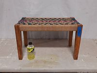 Small wooden bench with colorful woven seat on a plain floor with a yellow bottle in front for scale.
