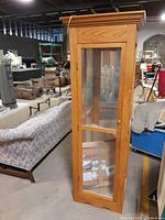 Front and side angled view of the tall wooden curio cabinet with glass panels, showing the natural wood finish, the crown molding, and scalloped base.