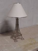 Front view of the Eiffel Tower design table lamp with white fabric lampshade on a plain surface with gray background.
