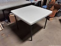 Angled front view of white plastic square folding table with metal legs on concrete floor.