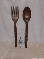 Photo of the large hand carved wooden fork and spoon set side by side with a water bottle for size reference showing decorative carved handles and smooth finish.