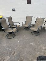 Four patio swivel chairs arranged on pavement next to a white wall. Chairs have metal frames with brown mesh seats and circular swivel bases.